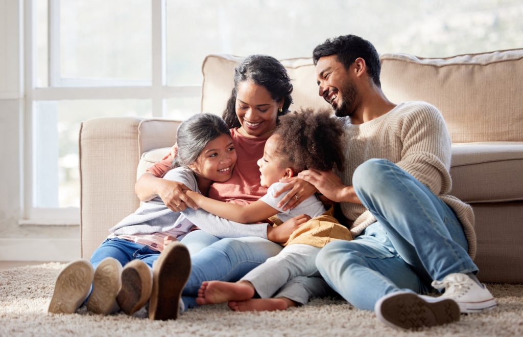 Une famille heureuse est assise sur le sol d'un salon lumineux. Les parents souriants sont entourés de leurs deux jeunes enfants, créant une atmosphère chaleureuse et aimante. La pièce est baignée de lumière naturelle provenant des grandes fenêtres, ajoutant à l'ambiance accueillante.